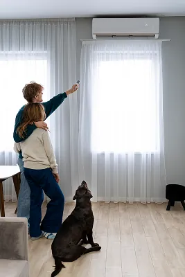 casal numa sala apontando controle remoto do ar condicionado, ao lado cachorro preto feliz olhando para o ar condicionado acima da janela.